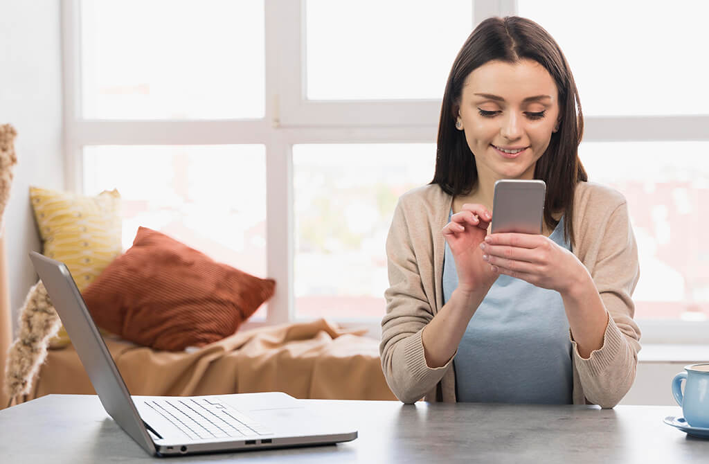Woman sitting at a desk using a smartphone with a laptop open beside her
